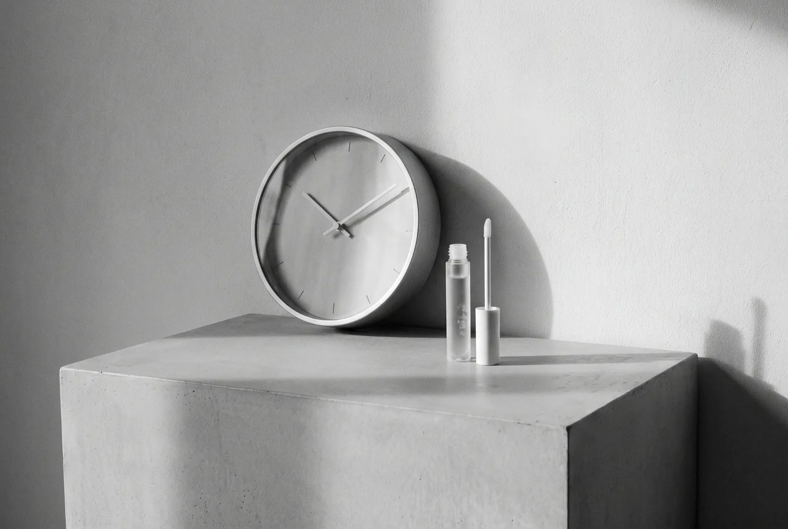 A minimalist wall clock and a brow serum tube on a grey plinth